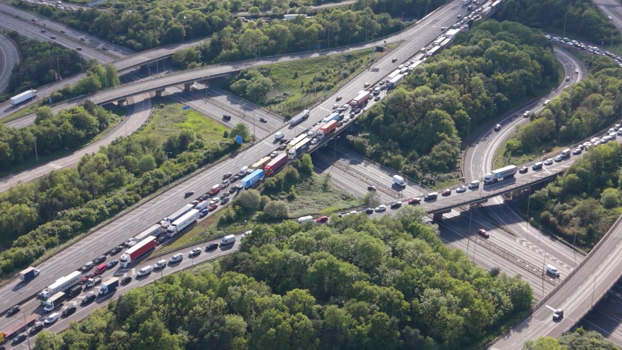 Eine Luftaufnahme zeigt tagsüber extreme Staus auf der Autobahn. Hunderte von Autos und Lastwagen stecken auf mehreren Fahrspuren eines belebten Autobahnkreuzes fest, umgeben von grünen Bäumen.