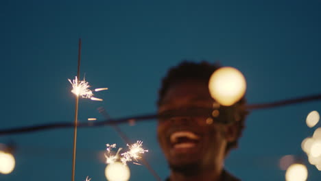 joven afroamericano sosteniendo bengalas bailando celebrando la víspera de año nuevo en la azotea por la noche divirtiéndose con amigos disfrutando de la celebración de fiestas navideñas