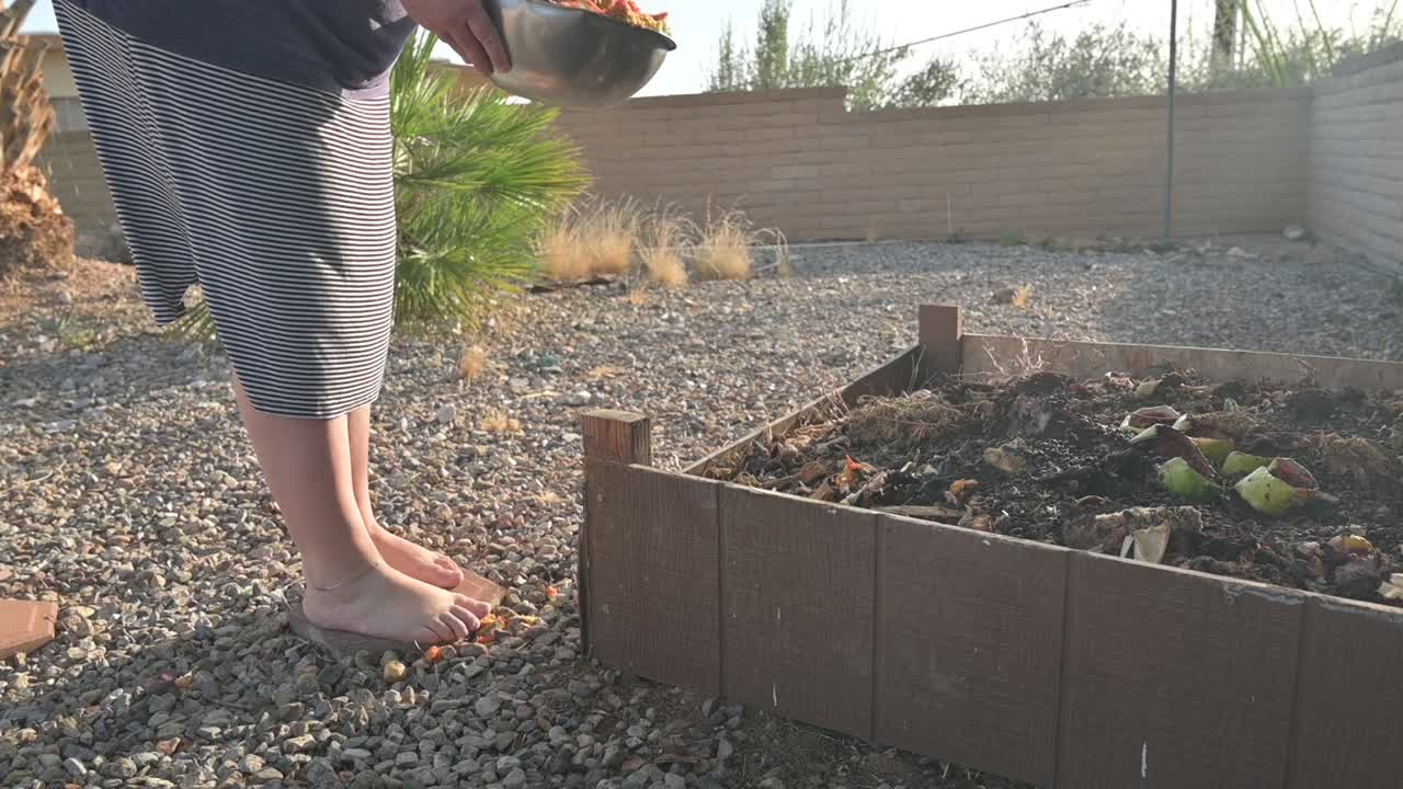 Woman approaches compost pile in backyard and empties out a bowl with organic fruit and vegetable scraps