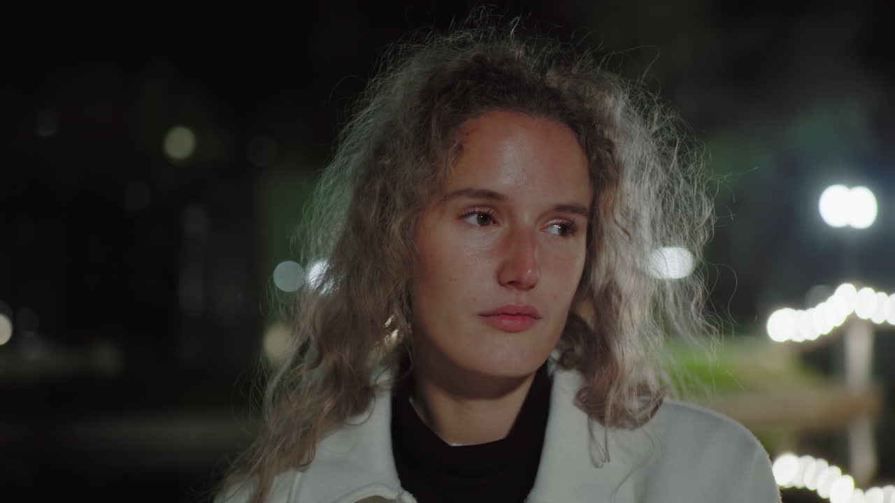 Closeup of thoughtful student alone at night, eyes closed in peaceful reflection, hair gently fluttering in wind, surrounded by softly glowing blurred streetlights in quiet urban atmosphere near water