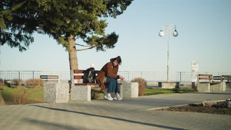 a girl is sitting alone on a bench in an urban park, looking visibly disconsolate. she is wearing a brown coat, blue jeans, and white shoes, with a black backpack beside her. deep contemplation