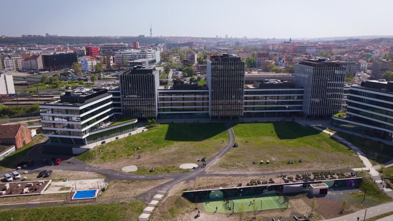 Filmische Luftaufnahme eines modernen Bürokomplexes und eines grünen Parks im Prager Stadtteil Holešovice mit Stadtsilhouette und dem Žižkov-Turm im Hintergrund an einem sonnigen Tag