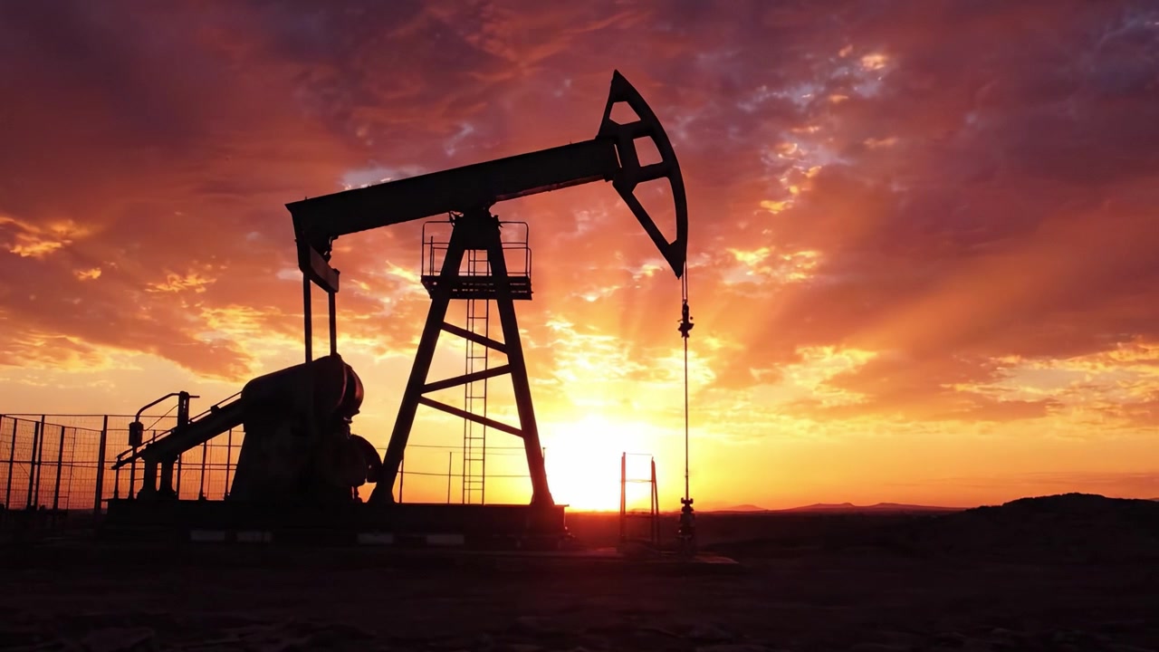 En betagende solnedgang over en oliepumpe, der tegner silhuet mod levende skyer, og fremhæver skæringspunktet mellem natur og industri i et slående landskab.