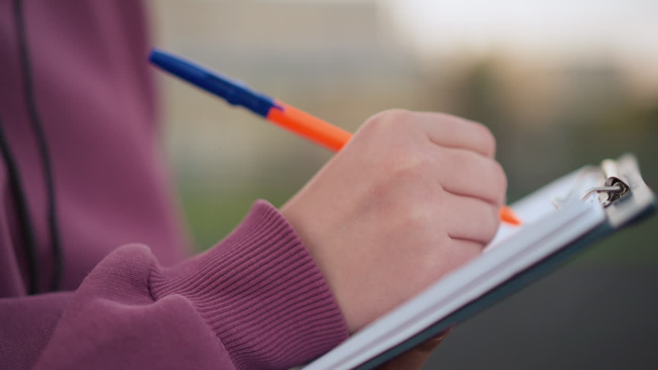 Close-up side view of individual in maroon top taking notes on clipboard outdoors, using an orange pen, background shows sports field with people walking