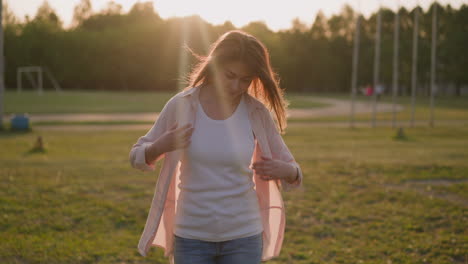 Smiling-woman-adjusts-shirt-walking-along-lawn-at-sunset