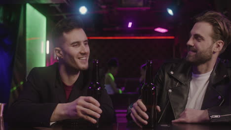 Two-Male-Friends-Toasting-And-Drinking-Beer-While-Sitting-At-Bar-Counter