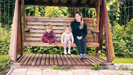 travelling young pretty mother with children swinging on a wooden swing near the summer house on vacation 1