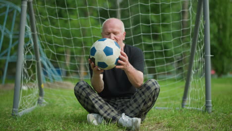 an elderly man sits cross-legged near a goal post, lifting a soccer ball that rolls towards him, he begins to observe the ball thoughtfully