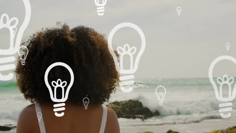 animation of light bulb icons over african american woman by seaside