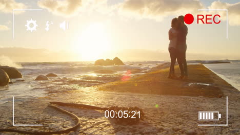 couple embracing on beach at sunset with camera recording animation overlay