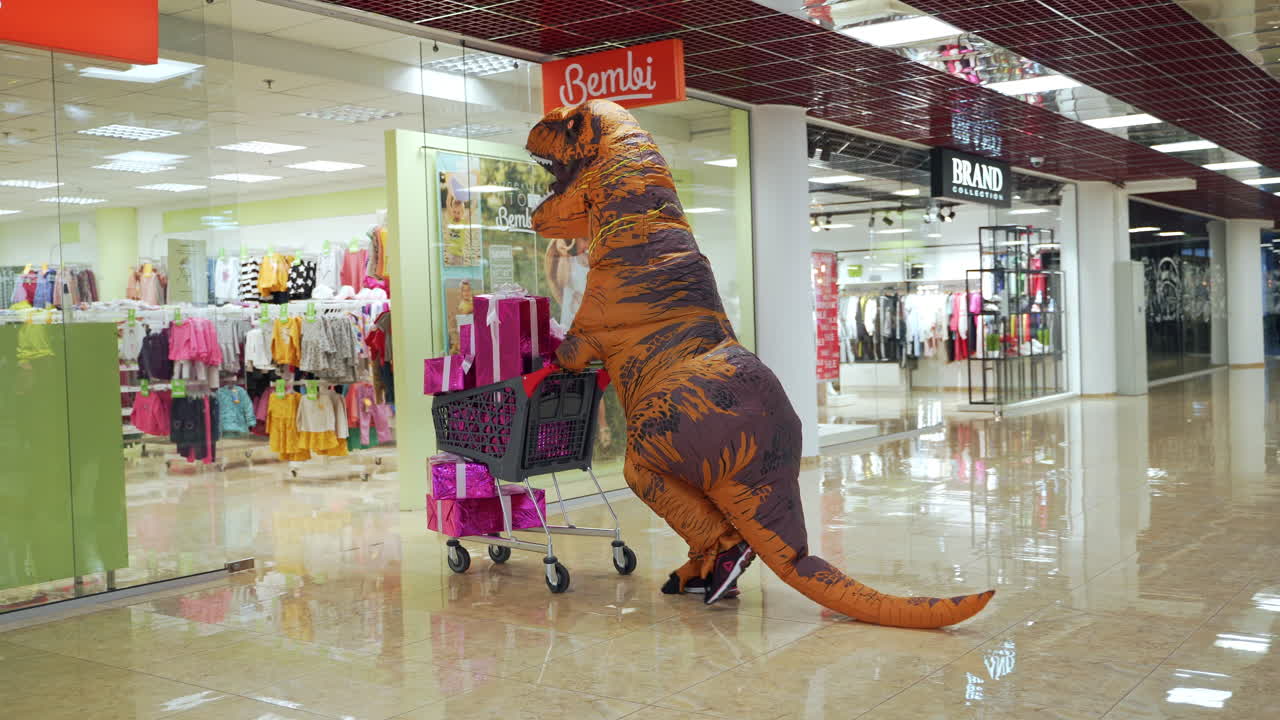 Dinosaur costumed man leaving the shop with a cart full of presents. Orange T-Rex enters the children's clothes store in shopping mall.