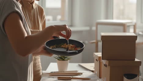 couple eating a meal from a pan while unpacking boxes