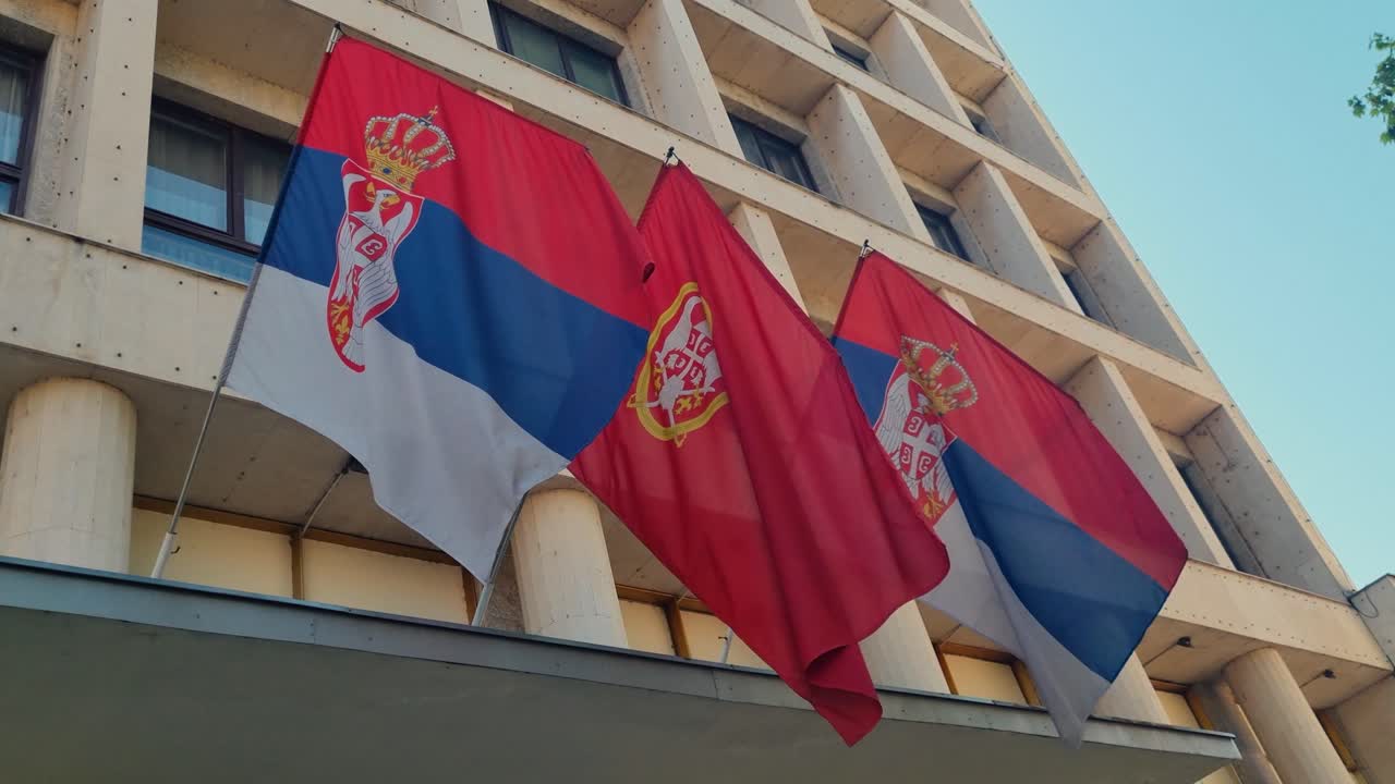 Serbian flags sway gently in a soft spring breeze, mounted on a Yugoslav-style government building under a clear blue sky
