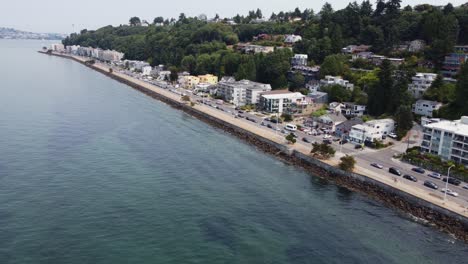 Drone-shot-showing-cars-driving-down-the-coast-at-Alki-Beach-in-Seattle,-Washington-with-the-Space-Needle-in-the-background