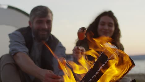 pareja alegre sentada junto a una carpa en la playa del lago y cocinando salchichas sobre un fuego para hacer un picnic en el campamento