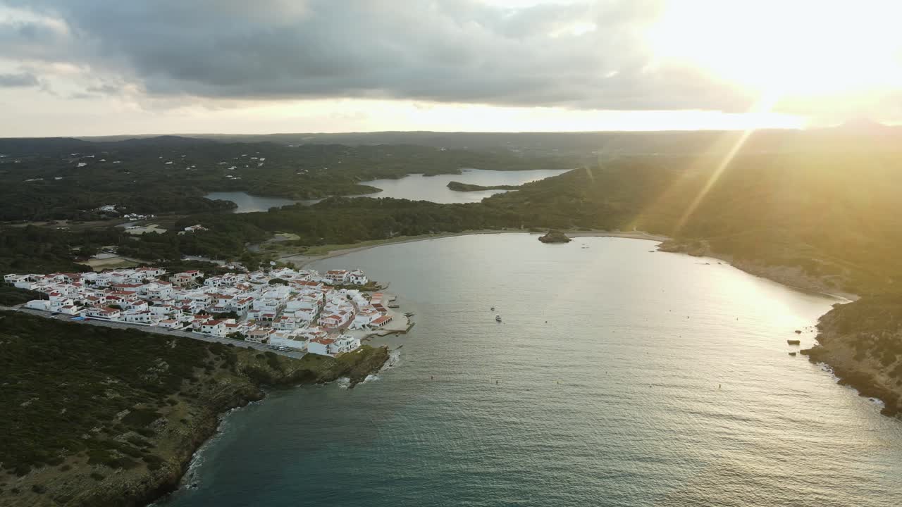 Sun rays shine down over the mountains to small village town of Es Grau in Menorca Spain. Drone cinematically panning around the harbour
