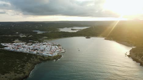 sun rays shine down over the mountains to small village town of es grau in menorca spain. drone cinematically panning around the harbour