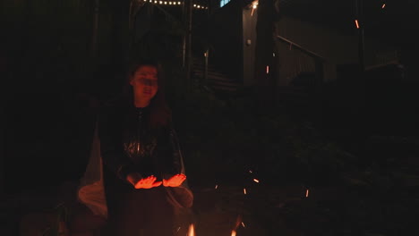 dama descansa en rocas ornamentales al lado de la fogata. joven novia pensativa calienta las manos sobre la hoguera mientras espera al chico en el patio trasero del campamento tarde en la noche