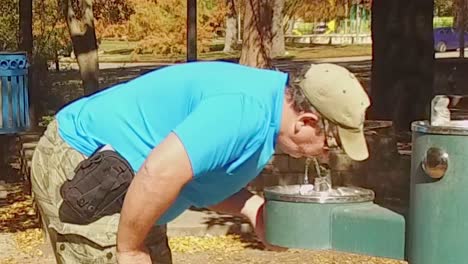 man drinking from drinking fountain in the park on a beautiful sunny day surrounded by trees and grass