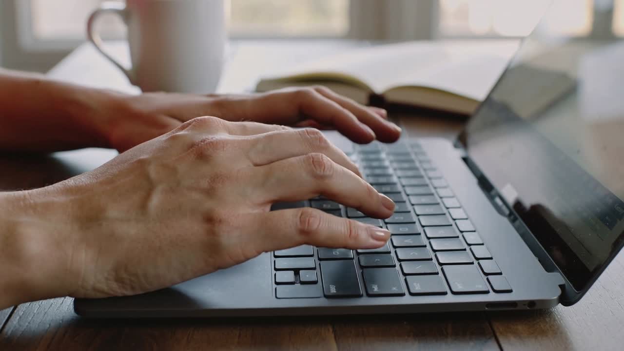 Dita che premono i tasti, mani adulte che digitano a una scrivania di legno, con laptop, tazza e taccuino.
