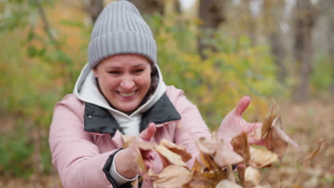 woman in pink jacket and grey beanie enjoys autumn, smiling as leaves fall around her poured by someone in the background in a vibrant display of seasonal joy