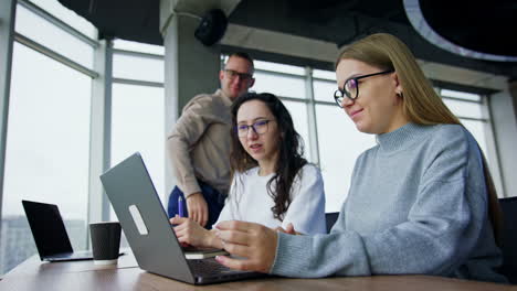young female colleagues look at laptop talking. male employee comes up suddenly sitting down on the desk. low angle view.