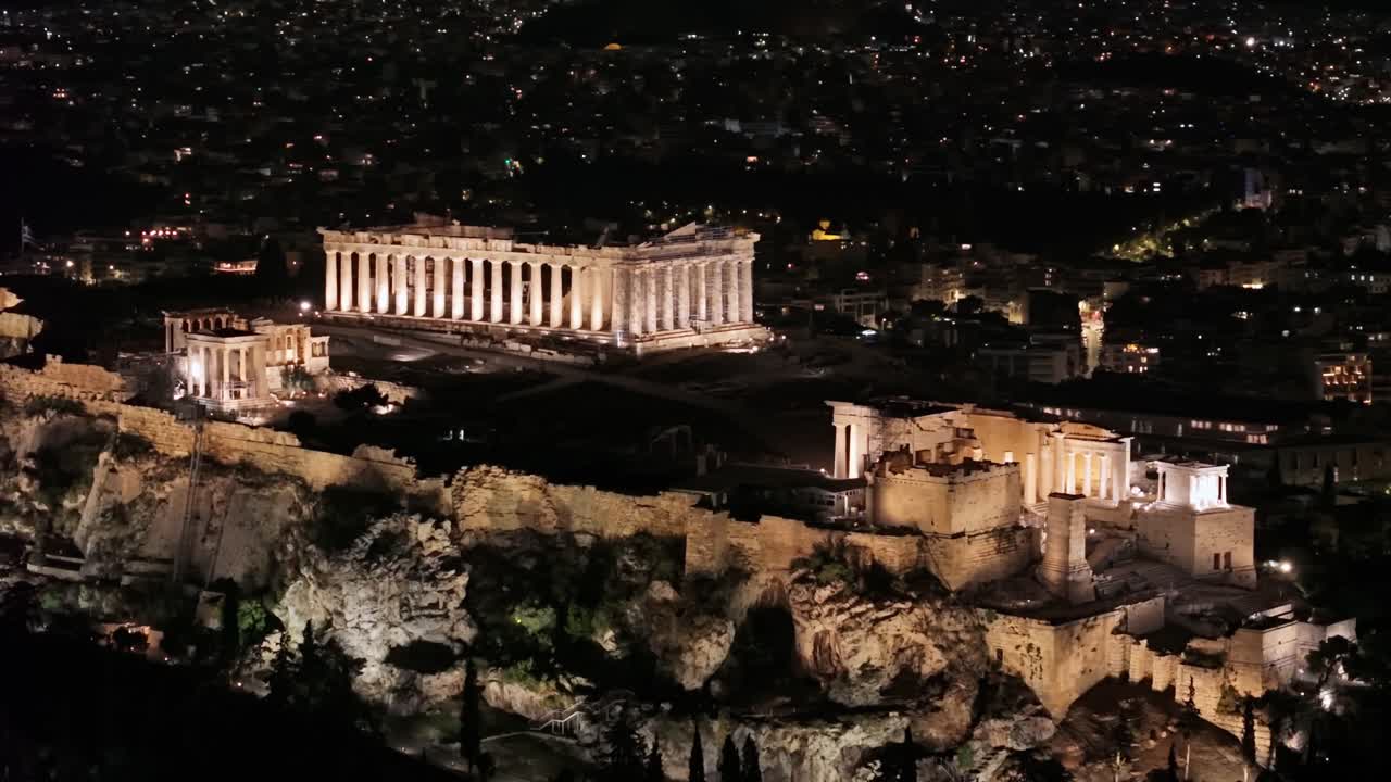 Ripresa orbitale di un drone mostra l'illuminata Acropoli e il Partenone di notte, con il panorama urbano di Atene che si estende sullo sfondo sotto il cielo scuro