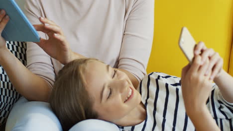 close up of blonde teenage girl scrolling and taping on smartphone while she is lying on her mother knees who is using tablet device