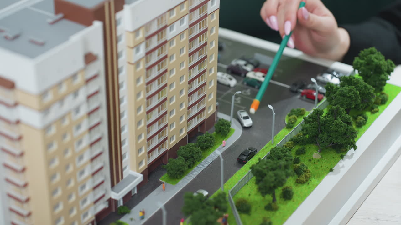 Close up hand view of realtor using pencil to explain architectural details of urban residential model with parked cars, miniature people, and surrounding greenery