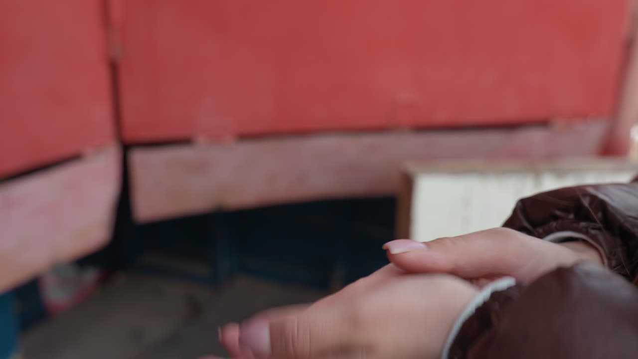 Primer plano de las manos de una mujer blanca frotándose cerca de una superficie metálica roja; se ven una chaqueta marrón y un puño; palmas y dedos en movimiento; gesto para calentar las manos en clima frío; detalle de la piel y la manga