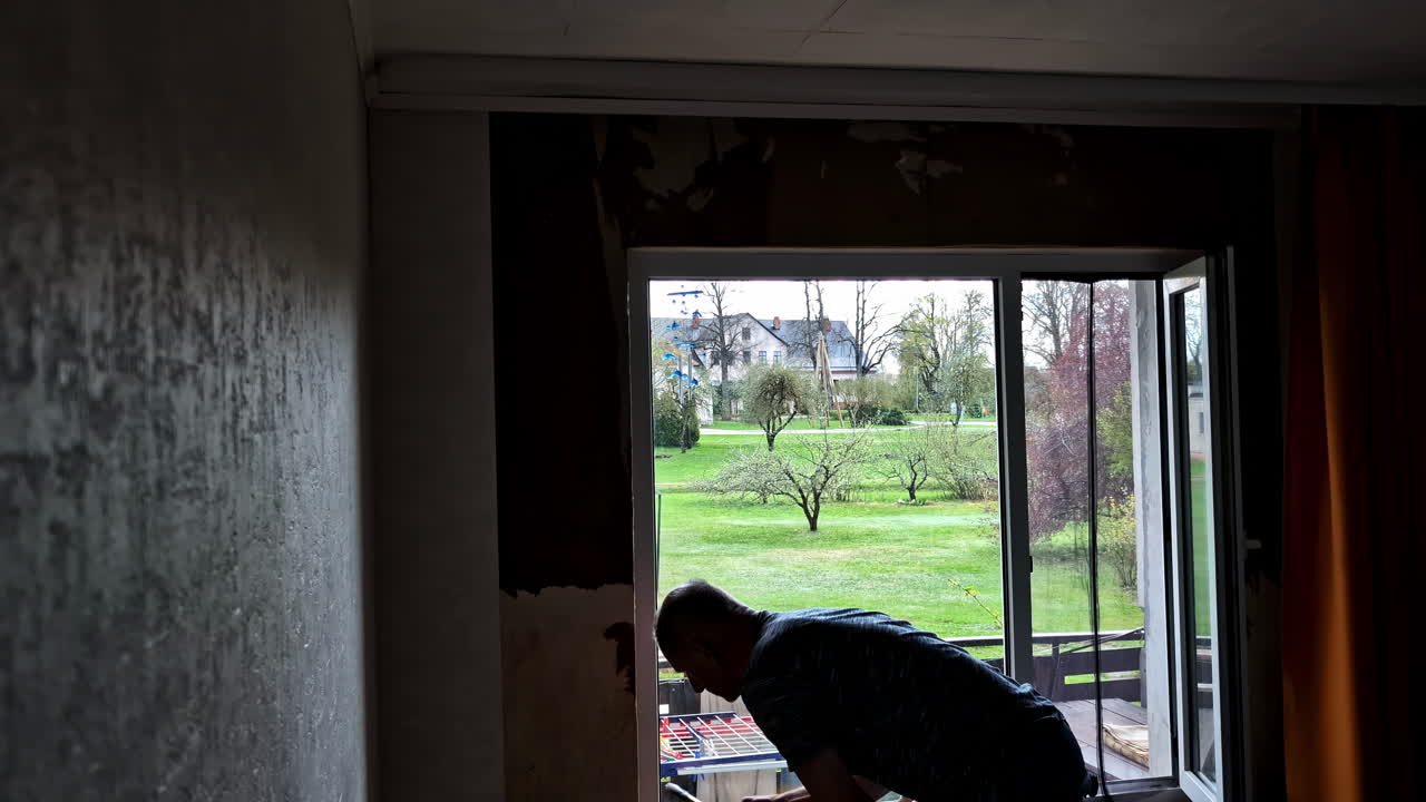 Man applying wallpaper adhesive to wall with brush in natural daylight