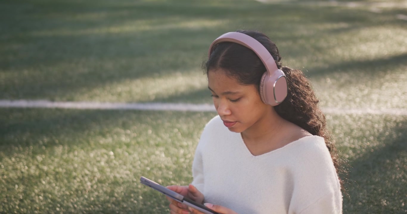 Bambina che raccoglie cuffie rosa su un campo sportivo, le indossa e tocca il tablet per giocare