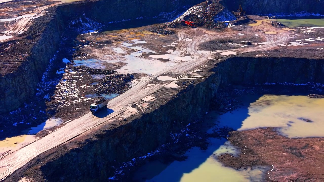 Drone shot of a dumper truck on a quarry road, Bulgaria. Elevated view of a white commercial vehicle driving along a steep dirt ridge near water and stone piles.