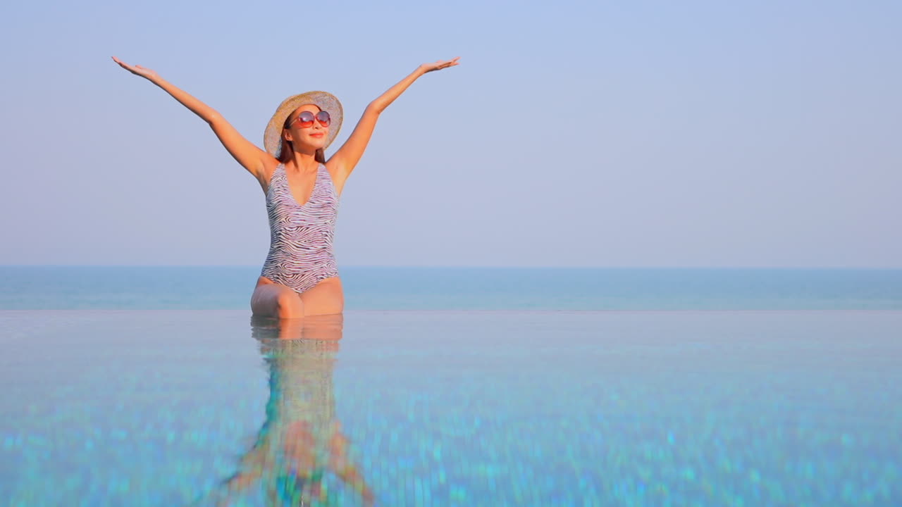 mientras se sienta en el borde de una piscina infinita, con el horizonte del océano al fondo, una mujer en traje de baño, sombrero para el sol y gafas de sol levanta los brazos con alegría