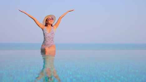 mientras se sienta en el borde de una piscina infinita, con el horizonte del océano al fondo, una mujer en traje de baño, sombrero para el sol y gafas de sol levanta los brazos con alegría