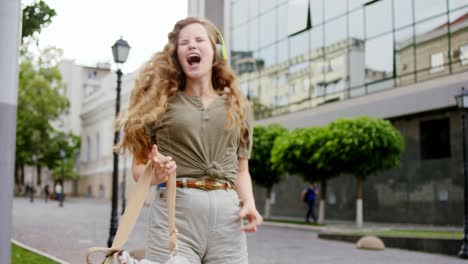 mujer joven divertida y carismática con cabello largo y rizado bailando en medio de la calle y disfrutando del tiempo mientras escucha música de los auriculares