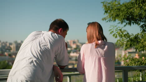 back view of boy and girl leaning over iron railing as they interact while girl shows something on a tablet, background features blurred cityscape and greenery