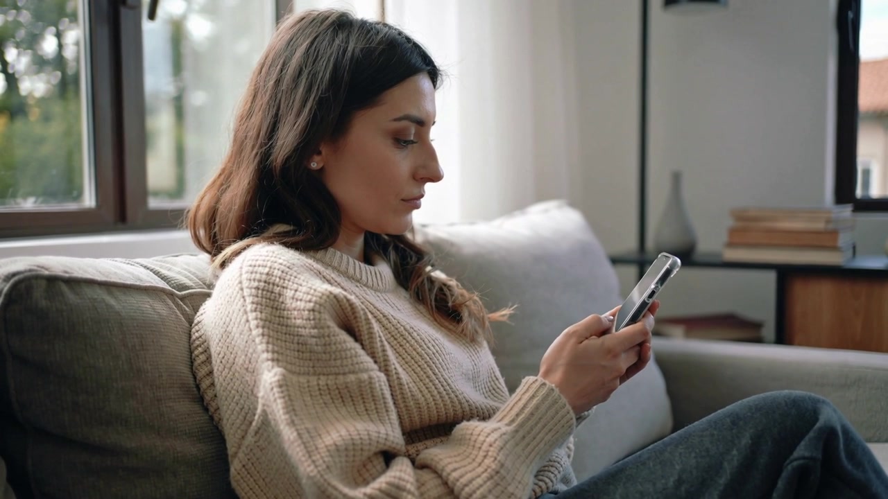 Dospělá žena v krémovém svetru zvedá smartphone, posouvá obsah a naklání hlavu na pohovce v obývacím pokoji.
