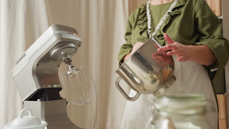 chef in green outfit playfully taps stainless bowl while dancing in kitchen, bowl reflects her moving hands, stand mixer and kitchen setup in view