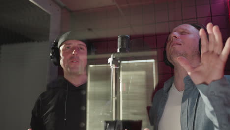 close-up of two men in a red soundproof room, singing passionately, fully immersed in the music. one wears a black cap and shirt, while the other is dressed in a blue shirt and white inner shirt