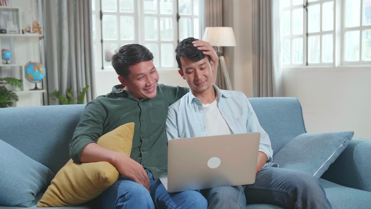 Asian Gentle Gay Couple Using Laptop Computer, While Sitting On A Couch In Cozy Stylish Apartment. Adult Boyfriends Online Shopping On Internet, Watching Funny Videos On Streaming Service