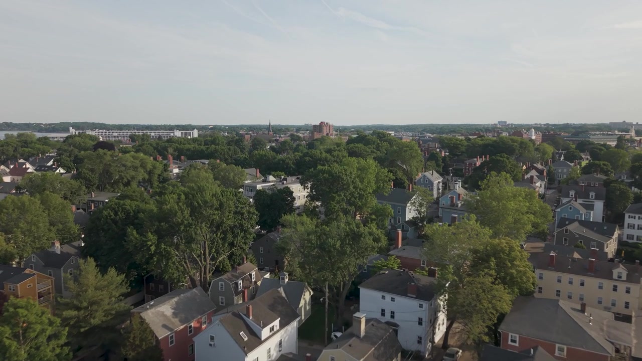 Case coloniali con camini in mattoni si annidano tra fitti alberi verdi, una lontana guglia di chiesa e l'acqua del porto si estendono verso lo skyline sfocato della città a Salem, Massachusetts, USA. Ripresa aerea con drone