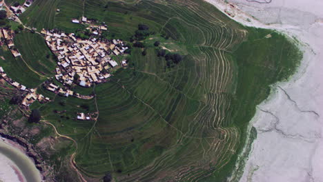 Aerial-rotational-view-over-the-village-surrounded-by-a-dry-river,-mountains-and-green-farms-and-trees