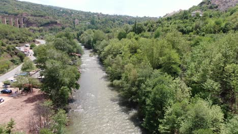 Drone-view-in-Albania-vertical-lift-over-rapids-river-between-green-mountains-and-a-big-column-bridge-on-a-sunny-day