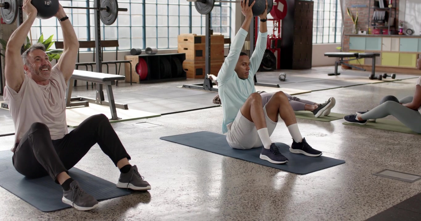 Diverse participants with center man performing 10lb ball sit-ups from supine on mats in gym