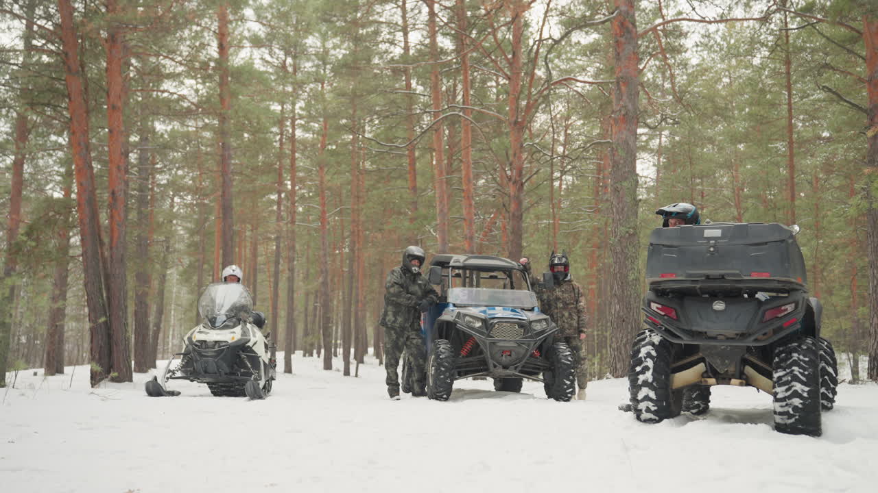 In una foresta di pini innevata, un convoglio di quad si prepara: la guida controlla il GPS, un meccanico ispeziona il motore, un pilota sale su una motoslitta accanto a un UTV. Alti pini, neve che cade dolcemente, piste ghiacciate e pneumatici robusti creano un'atmosfera di spedizione e lavoro di squadra.