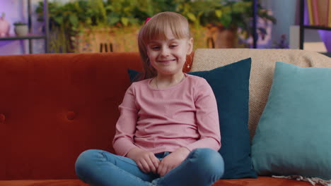 Cheerful-lovely-caucasian-little-toddler-kid-girl-smiling,-looking-at-camera-sitting-on-sofa-at-home
