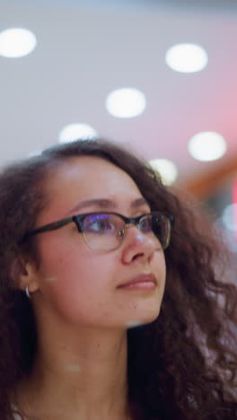 mujer con gafas y cabello rizado mirando a su alrededor en un centro comercial bien iluminado, iluminado por suaves luces bokeh en el fondo y la gente caminando en el centro comercial borroso