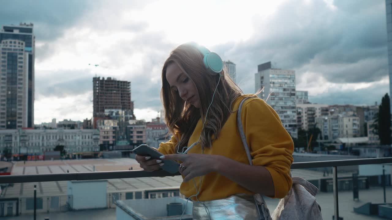 Portrait of young cute attractive young girl in urban city streets background listening to music with headphones. Woman wearing yellow blouse and silver skirt