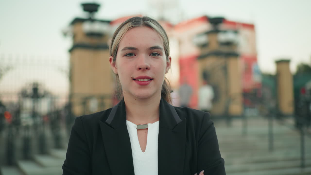Confident woman with folded arms standing outdoors with elegant smile, dressed in business attire, exuding calm assurance and professionalism, in urban setting with blurred